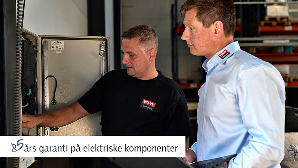 To VELUX Commercial-teknikere inspicerer et åbent elektrisk kontrolpanel i et industrielt miljø. Tekst på billedet: "25 års garanti på elektriske komponenter."