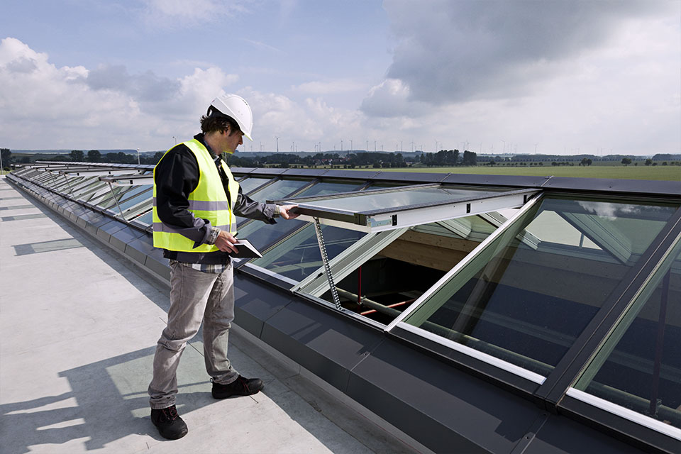 A service instructor wearing a safety helmet and high-visibility vest is inspecting a series of VELUX Modular Skylights on a large, flat rooftop