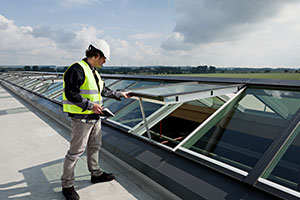A service instructor wearing a safety helmet and high-visibility vest is inspecting a series of VELUX Modular Skylights on a large, flat rooftop