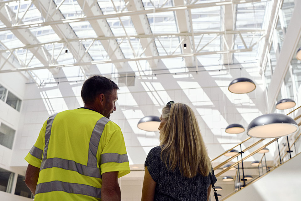 A service instructor in a reflective safety vest is speaking with a customer as they both look up at the rooflight inside a bright, modern atrium
